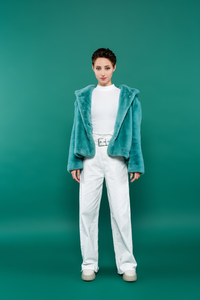 full length view of brunette woman in faux fur jacket and white trousers looking at camera on green - Foto, afbeelding