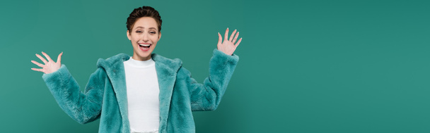 ενθουσιασμένοι γυναίκα σε faux γούνα σακάκι κουνώντας τα χέρια, ενώ κοιτάζοντας κάμερα που απομονώνονται στο πράσινο, πανό - Φωτογραφία, εικόνα