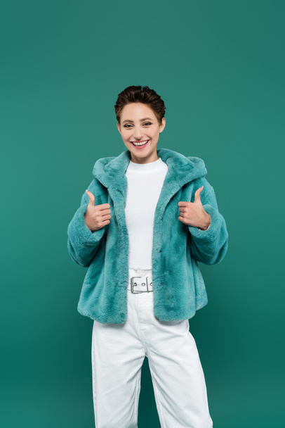 cheerful woman in trendy faux fur jacket showing thumbs up isolated on green - Fotografie, Obrázek