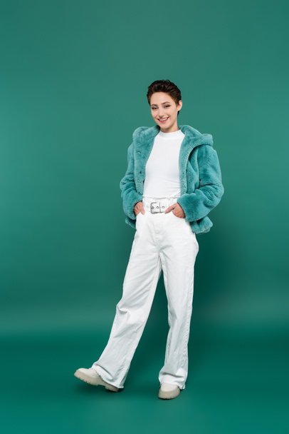 happy and stylish woman in faux fur jacket posing with hands in pockets of white trousers on green - Фото, изображение