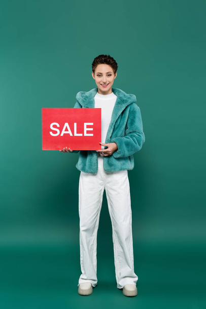 full length view of happy woman in white trousers and faux fur jacket holding red card with sale lettering on green - Zdjęcie, obraz