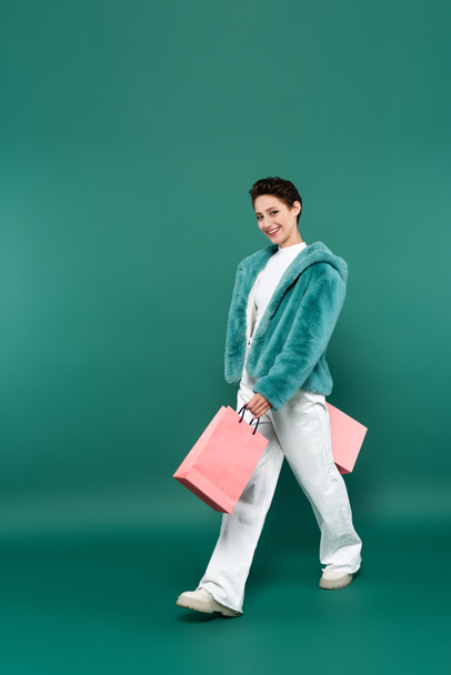 femme joyeuse en fausse veste de fourrure et pantalon blanc marchant avec des sacs à provisions roses sur vert - Photo, image