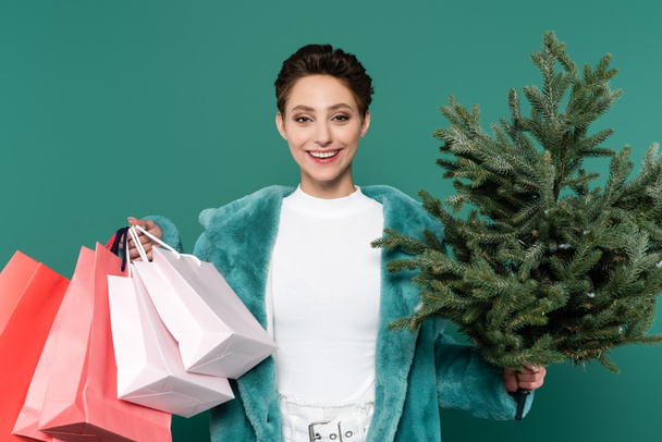 Alışveriş torbaları ve küçük köknar ağacı olan mutlu ve şık bir kadın yeşile izole edilmiş kameraya bakıyor. - Fotoğraf, Görsel