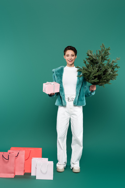 vista completa de la mujer complacida con caja de regalo y pequeño pino cerca de bolsas de compras en verde - Foto, Imagen
