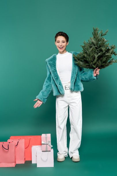 full length άποψη του ευτυχισμένη και μοντέρνα γυναίκα με μικρό χριστουγεννιάτικο δέντρο δείχνοντας τσάντες ψώνια στο πράσινο - Φωτογραφία, εικόνα