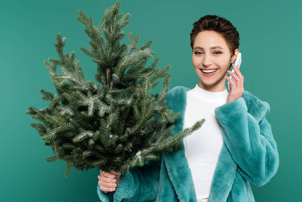 fashionable woman with small fir tree smiling while talking on mobile phone isolated on green - Photo, Image