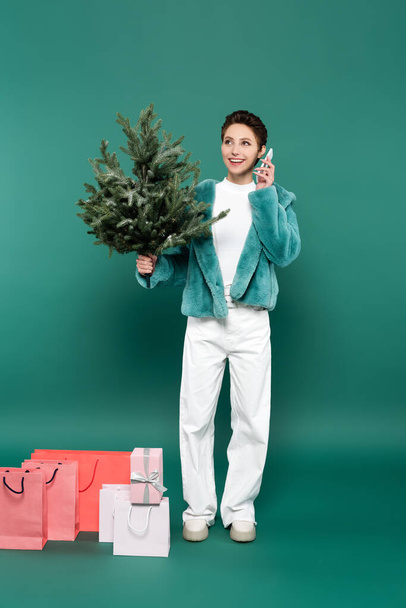trendy and happy woman with small fir tree talking on mobile phone near shopping bags on green - Photo, Image