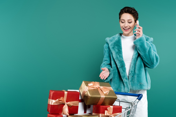 stylish woman smiling while talking on smartphone and pointing at gift boxes in shopping trolley isolated on green - Photo, Image