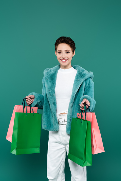mujer feliz y de moda con bolsas de compras multicolores sonriendo a la cámara aislada en verde - Foto, Imagen