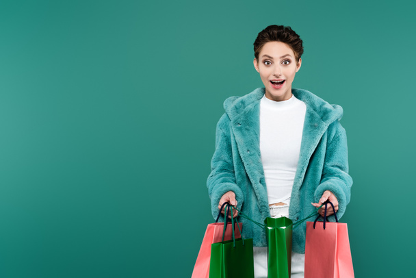 изумленная брюнетка в искусственном меховом пиджаке, держащая сумки с покупками, глядя на камеру, изолированную на зеленый - Фото, изображение