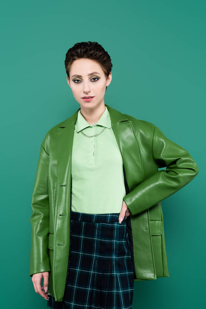 young woman in leather jacket and plaid skirt posing with hand on hip isolated on green - Foto, imagen