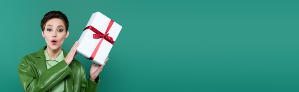 amazed brunette woman looking at camera while holding white gift box isolated on green, banner - Foto, immagini