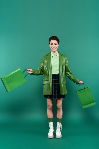 full length view of smiling woman in leather jacket, plaid skirt and white boots posing with shopping bags on green - Fotoğraf, Görsel