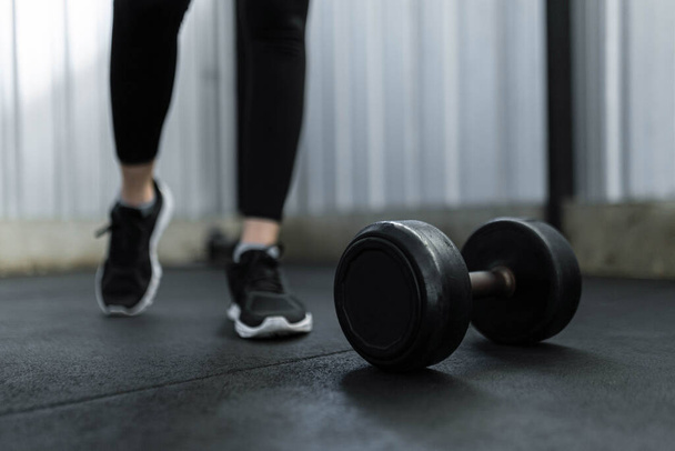 exercise concept The black rubber coated round dumbbell lying on the gym ground in front of the person who wears black trainers. - Foto, afbeelding