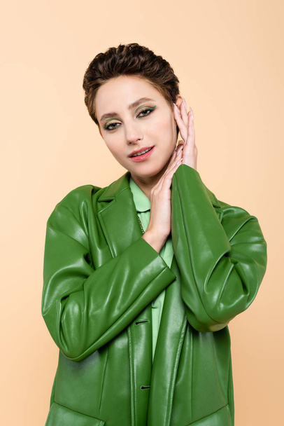 pretty brunette woman in trendy green jacket posing with hands near face isolated on beige - Foto, immagini