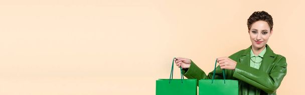 positive brunette woman in stylish jacket holding green shopping bags isolated on beige, banner - Foto, afbeelding