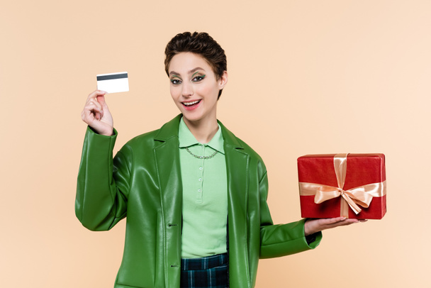 mujer feliz en chaqueta verde de pie con tarjeta de crédito y caja de regalo roja aislada en beige - Foto, Imagen