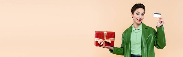 trendi barna nő piros ajándék doboz és hitelkártya mosolygós kamera elszigetelt bézs, banner - Fotó, kép