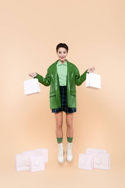 full length view of excited woman in trendy clothes levitating with shopping bags on beige - Foto, Imagen