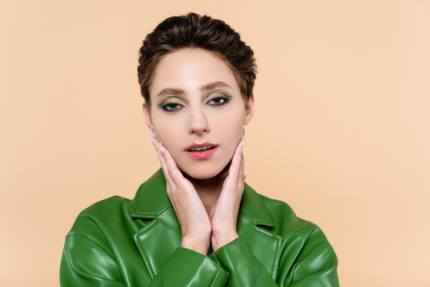 brunette woman in green leather jacket touching face while looking at camera isolated on beige - Φωτογραφία, εικόνα