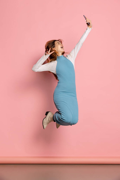 Cheerful young beautiful girl wearing warm blue dress jumping and using phone isolated on pink color background. Emotions concept - Valokuva, kuva