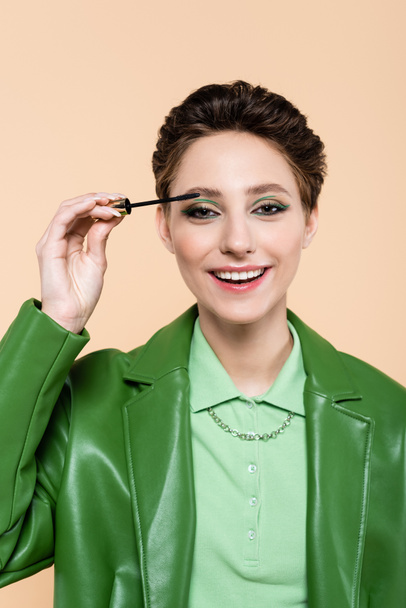 happy and stylish woman applying mascara while looking at camera isolated on beige - Photo, image