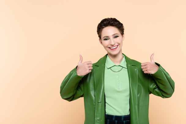 joyful woman in green stylish jacket showing thumbs up isolated on beige - Фото, изображение