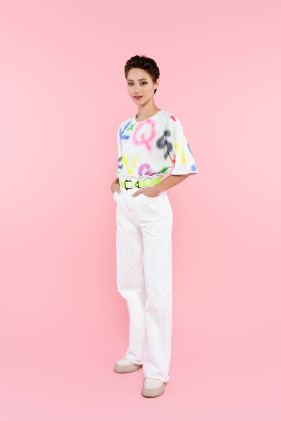full length view of stylish smiling woman standing with hands in pockets of white trousers on pink - Фото, зображення