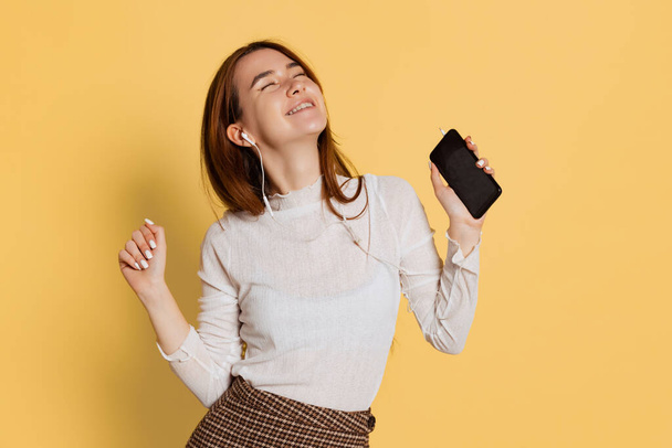 Cheerful young beautiful girl wearing warm white neck-polo in earphones using phone isolated on yellow color background. Emotions concept - Foto, Imagem