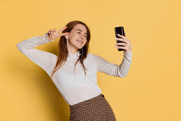 Cheerful young beautiful girl wearing warm white neck-polo in earphones using phone isolated on yellow color background. Emotions concept - Fotografie, Obrázek