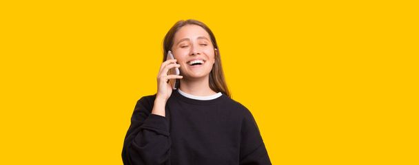 opgewonden jonge vrouw lachen als ze chats op een mobiele telefoon over gele achtergrond - Foto, afbeelding