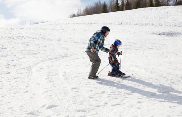 παιδί με τον μπαμπά μαθαίνει σκι κατά τη διάρκεια των χειμερινών διακοπών σε χιονισμένα βουνά την ηλιόλουστη κρύα ημέρα. Χειμωνιάτικες βόλτες με παιδιά. Εποχιακές χαρές, ευτυχισμένη παιδική ηλικία. Αθλητική εκπαίδευση, υποστήριξη, ομαδικότητα - Φωτογραφία, εικόνα