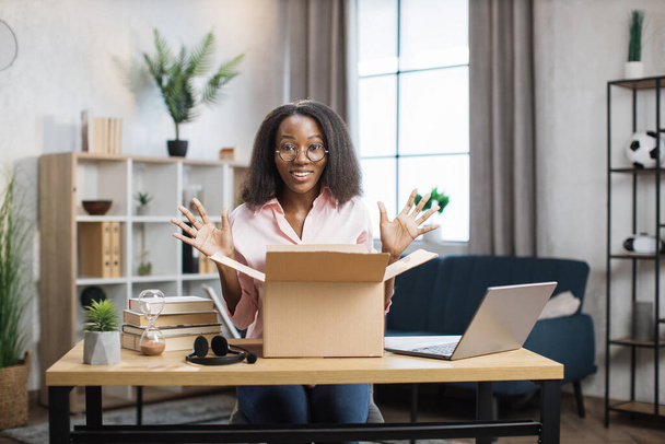 Excited african woman sitting at desk and opening parcel box - Foto, afbeelding