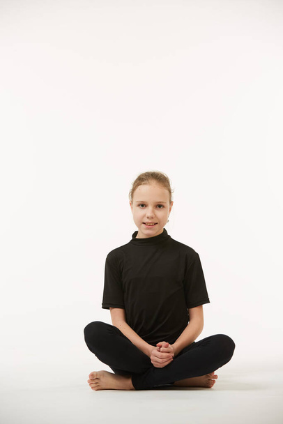 Una niña en edad escolar vestida de negro está parada sobre un fondo blanco y limpio. Movimientos de baile o calentamiento deportivo. - Foto, Imagen