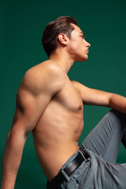 Profile view of young handsome sportive shirtless man wearing jeans sitting on floor isolated on green studio background. - Photo, Image