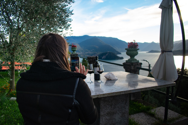 Jolie touriste féminine prenant des photos avec son téléphone du lac de Côme (Alpes italiennes) tout en buvant un verre de vin à la maison de vacances. - Photo, image