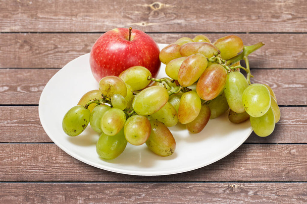 Ein Bündel grüner Trauben und ein Apfel auf einem Teller mit dem hölzernen Hintergrund. Ansicht von oben. - Foto, Bild