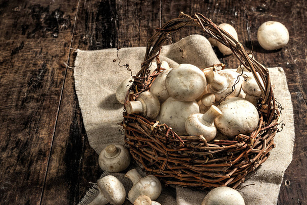 Fresh champignons in a basket. Raw ingredient for cooking vegan food. Old wooden background, fashion hard light, dark shadow, rustic style, copy space - Photo, Image