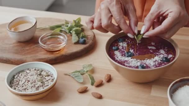 Close up tracking shot of unrecognizable young woman adding mint into healthy smoothie bowl. Nuts, sesame and pumpkin seeds, coconut flakes, blueberries and honey in bowls on table - Footage, Video