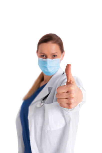 Προσωπογραφία νεαρής γυναίκας γιατρού με ιατρική μάσκα που κοιτάζει την κάμερα και δείχνει τον αντίχειρα προς τα πάνω, απομονωμένη σε λευκό φόντο. Ικανοποιημένος γιατρός με λευκό παλτό καλά αποτελέσματα στη μελέτη του ιού, ανακαλύψτε το εμβόλιο - Φωτογραφία, εικόνα