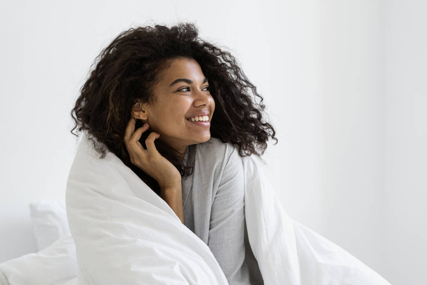 幸せな暗い肌の女性は柔らかい毛布に包まれ、脇を見て、ベッドルームで早朝を過ごす。夢遊病の週末を家で楽しみながら目を覚まし - 写真・画像