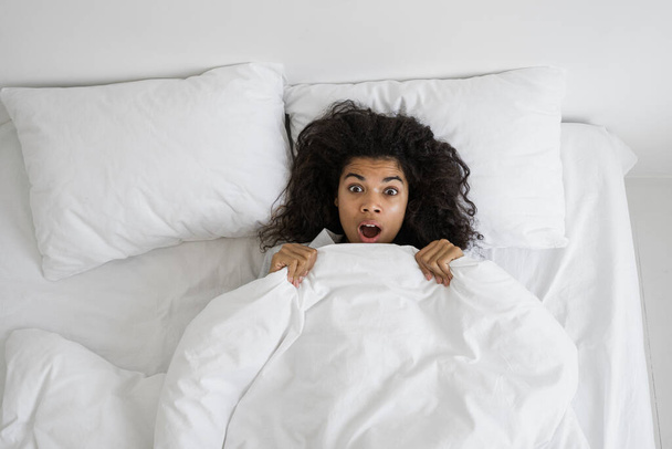 Nightmare or bad dream concept. Above view of stressed afro american woman with shocked face looking at camera. Scared female in panic lying on bed in sleepwear, screaming after sleep - Photo, Image