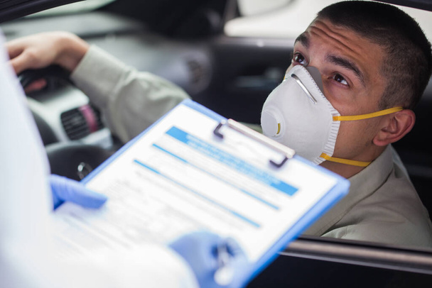 Closeup of doctor or healthcare worker in protective medical gloves writing down patient info in clipboard,young caucasian man wearing protective face mask in mobile car testing or vaccination center - Фото, изображение