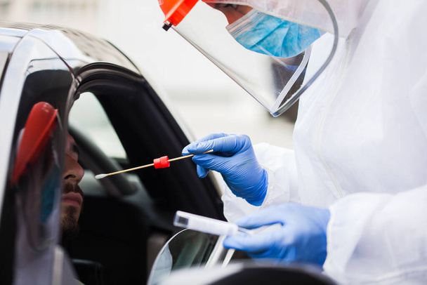 Medical worker wearing protective PPE surgical gloves and clothing,holding nasal swab kit,performing drive-thru COVID-19 testing,point of care on site location PCR detection of new Coronavirus variant - Fotografie, Obrázek