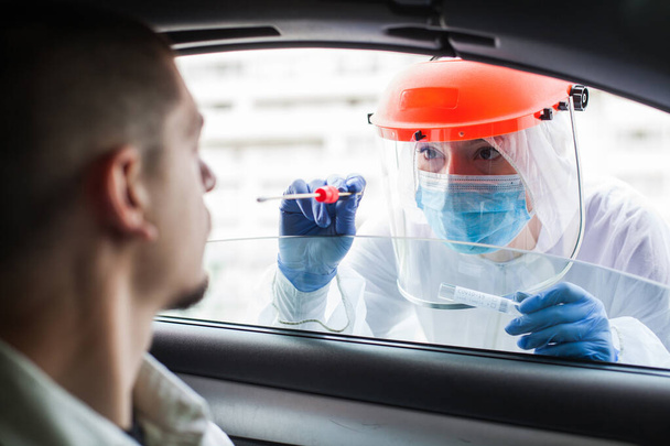 Female UK medical technician in full protective personal protective equipment gear collects swab sample from young man sitting inside car,Coronavirus mobile testing drive-thru center,COVID-19 pandemic - 写真・画像