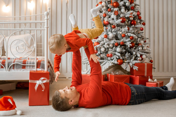 Happy young father lying on the floor near the New Year tree and a lot of present boxes at christmas time and holding his funny little son in front of him in his stretched arms - Фото, изображение
