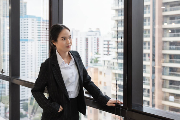 Joven mujer de negocios asiática de pie y mirando por la ventana en el edificio de oficinas en el centro - Foto, imagen