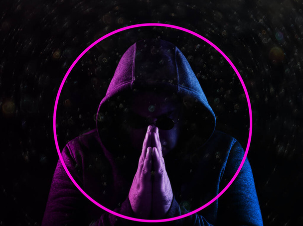 Portrait of fashion man in a hoodie with a hood and round sunglasses in neon light. studio shot. Holding hands folded in prayer.  isolated on black background. - Фото, зображення