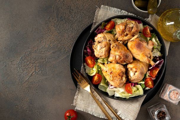 Fried chicken legs with salad in a plate on a dark kitchen table. Cooked poultry meat, pickles, and tomatoes on a culinary background top view - Photo, image