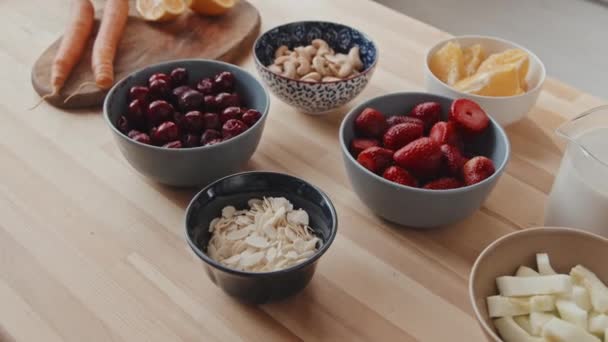 Ruční zblízka záběr čerstvých ingrediencí na kuchyňském stole bobule, ovoce a ořechy v miskách, mléko v džbánu a loupaná mrkev na dřevěné řezací desce - Záběry, video